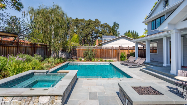 A beautiful inground pool with a stone deck surrounded by a wood fence with a jacuzzi made of stone.