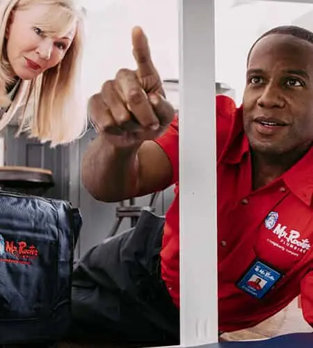 Mr. Rooter plumber pointing out something under a sink to female customer.