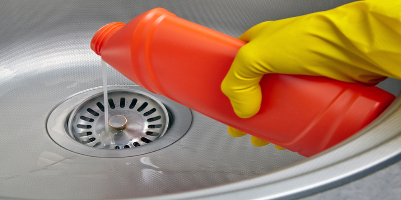 Hand in yellow rubber glove pour sewer pipe cleaner down the kitchen sink drain.