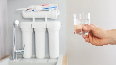 A man holding a glass of water with a reverse osmosis system on a kitchen counter next to a sink in the background.