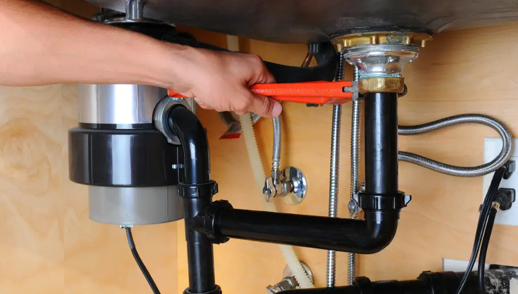 A plumber using a wrench to complete garbage disposal installation underneath a sink in a residential kitchen.