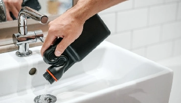 A man’s hand pours a chemical drain cleaner from a black bottle into a white sink.