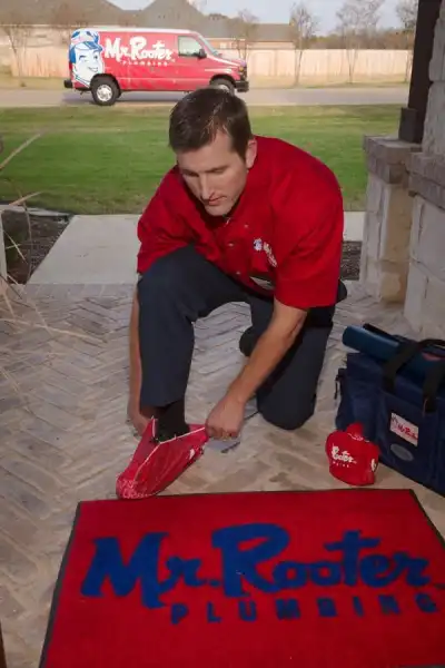 Mr. Rooter plumber putting on shoe covers at client's door, with Mr. Rooter mat in front of him and Mr. Rooter van parked on street behind him.