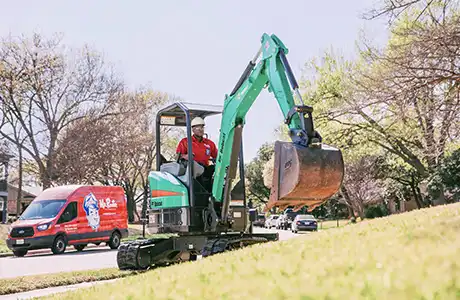 Mr. Rooter technician driving an excavator in a client's front yard, with branded company van parked on street behind him.