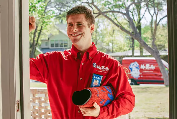 Smiling Mr. Rooter plumber knocking at customer's front door, holding rolled-up mat, with company van parked on street behind him.